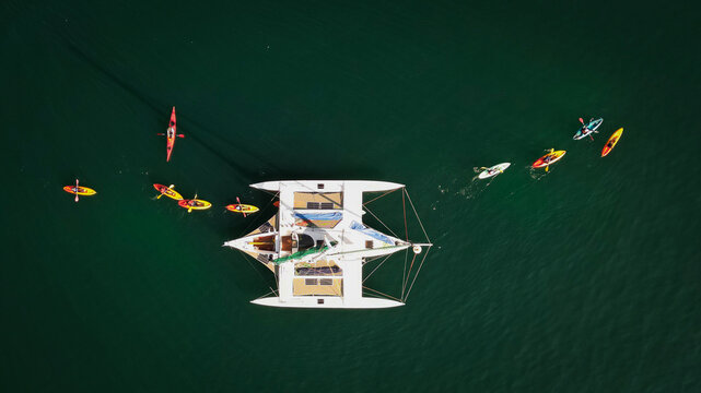 Aerial view of a white catamaran surrounded by colorful kayaks in the clear water near Pointe du Cosmeur in Carantec, Bretagne, France.