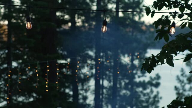 Warm string lights glow through smoky forest beside calm lake, creating a cozy campsite atmosphere with soft bokeh, dark trees and evening haze above the water