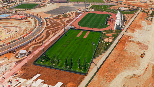 Aerial view of a sports complex under construction featuring green fields, a running track, and stadium lighting surrounded by dirt and ongoing development in Athens, Greece.