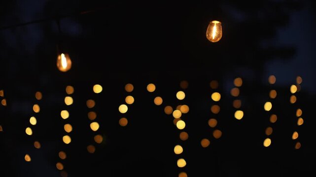 Warm glowing bulbs and blurred fairy lights decorate dark forest, creating cozy nighttime campsite illumination with golden bokeh against deep blue and black woodland shadows