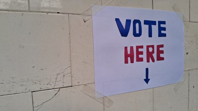 Handwritten vote here sign taped to a white tiled wall surface