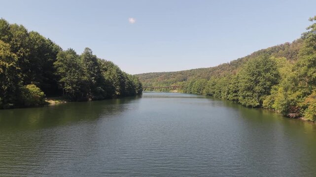 Stunning aerial perspective flying over a tranquil river in Germany, surrounded by a lush green forest, moving toward the historic Tatzlwurm bridge