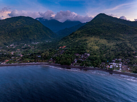 Aerial view of the Amed landscape featuring lush green mountains meeting the dark volcanic sand beach along the coastline of the Bali Sea at dusk, Abang, Bali, Indonesia.