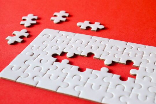 High angle perspective of an incomplete white jigsaw puzzle showing a rectangular gap over a solid red textured background.