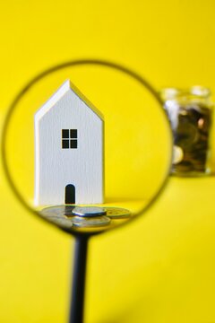 White wooden house model viewed closely through a magnifying glass with a blurred coin jar in the background.
