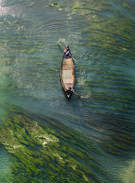 Aerial view of a wooden boat with two people rowing through green aquatic plants in a river in Sirajganj, Rajshahi Division, Bangladesh.