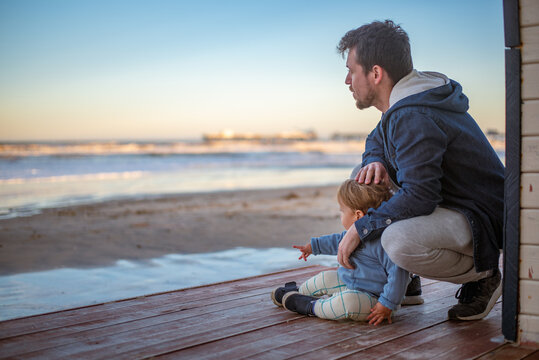 A 20-month-old toddler boy standing on the beach with his father and pointing at something in the distance