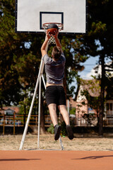 Basketball player performs a slam dunk at an outdoor court on a sunny day with clear skies and trees in the background © Nikola Spasenoski