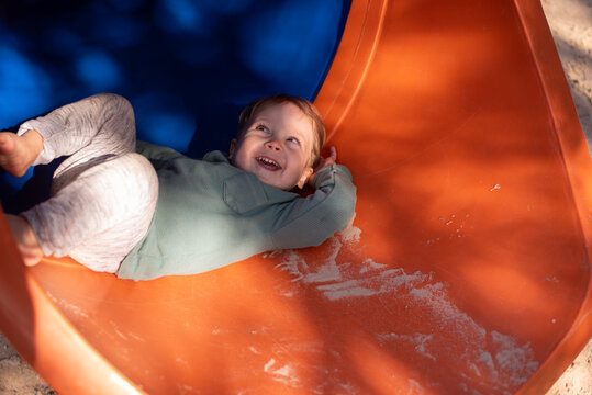 A happy 20-month-old toddler boy smiling as he goes down a covered playground slide at a sandy playground