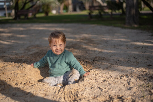 A 20-month-old toddler boy sitting on the sand with a gentle smile, fully dressed and enjoying a quiet outdoor moment