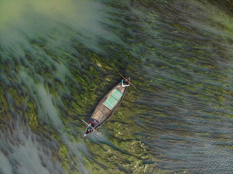 Aerial view of a small wooden boat with a person rowing on a river filled with green aquatic plants in Sirajganj, Rajshahi Division, Bangladesh.