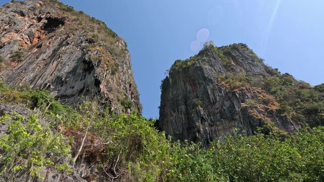 Wide shot of two massive limestone cliffs covered with dense greenery, surrounded by jungle and a beach in a nature reserve in Thailand under bright midday sun.