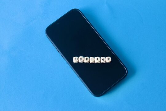 An overhead shot of a black smartphone centered on a clean blue background with small wooden cubes spelling out the word OFFLINE.