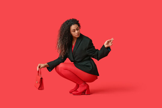 Beautiful young African-American woman in stylish red tights with handbag sitting on color background