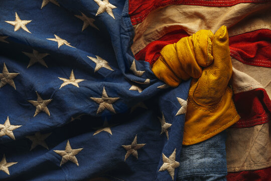 Hand in worn work glove holding the American flag. Symbolic of hardworking blue collar workers, USA manufacturing, pride in products Made in United States of America, or to celebrate US Labor Day.