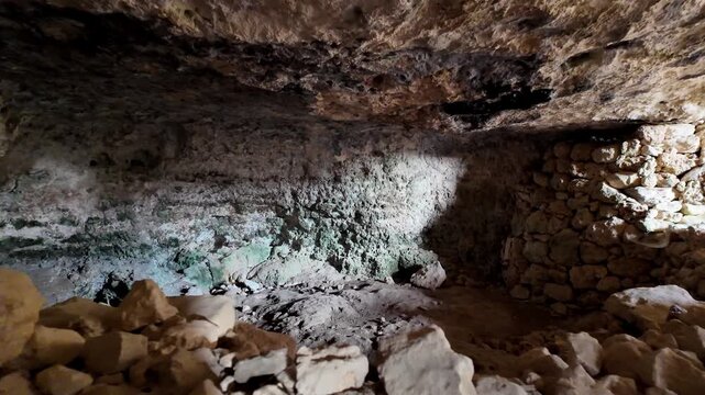 Ancient cave walls show weathered stone and green mineral streaks. Rocks and rubble fill the dim, mysterious chamber. Light spills through a narrow opening, casting shadows