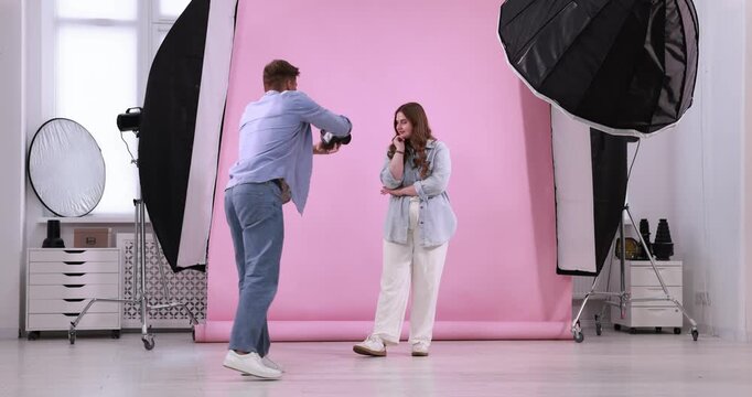 Plus size model posing for photographer with camera against pink background in studio. Beautiful young overweight woman