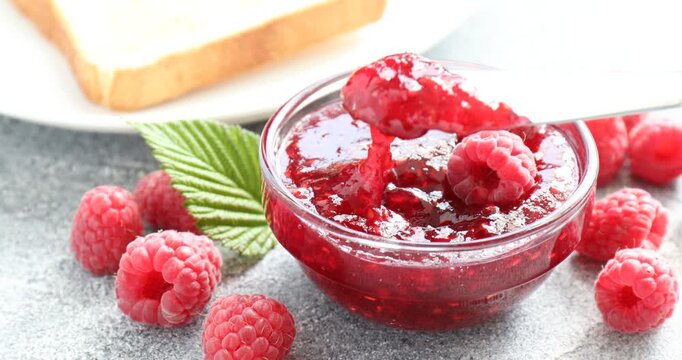 Putting tasty raspberry jam onto toast on grey table, closeup