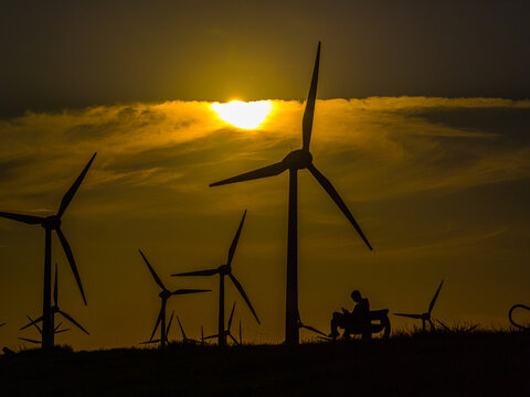 Aerial view of wind turbines silhouetted against a vibrant golden sunset with a person sitting on a bench in the foreground in Emden, Niedersachsen, Germany.