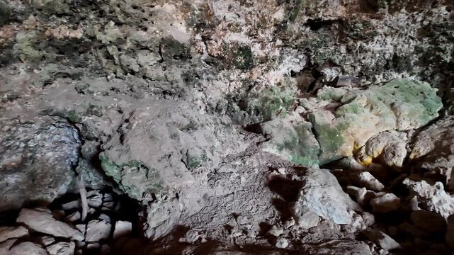 Dark cave with rough rock walls and scattered stones. Mossy surfaces glow under dim, artificial light. Water flows quietly over uneven terrain. Cave interior feels ancient and mysterious