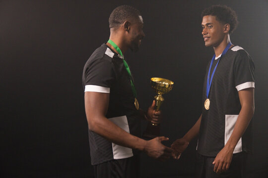 African male athletes exchanging gold trophy on stage, wearing matching black shirts and medals