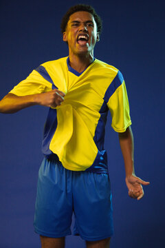 African American man pulling yellow blue soccer kit, shouting in studio lighting with blue backdrop