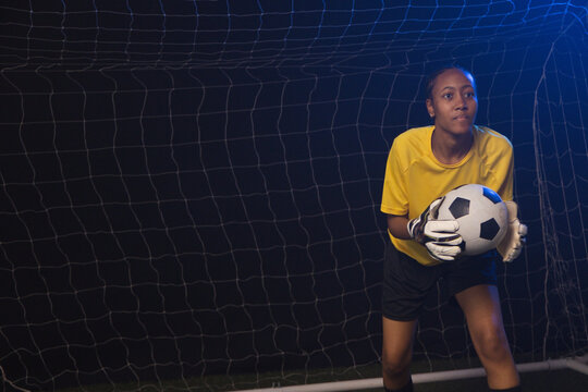 African American woman keeper holding soccer ball in gloves, yellow jersey by goal net, copy space