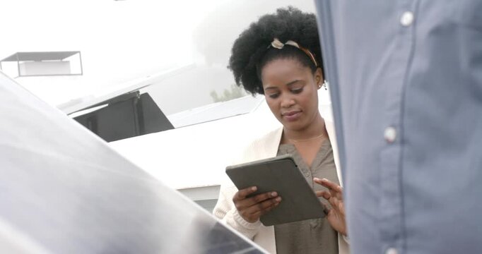 Coworker adjusting solar panel as African American woman holding tablet, tapping, logging roof data