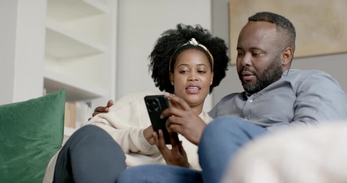African American couple on sofa at home, woman holding phone, tapping, showing, man smiling
