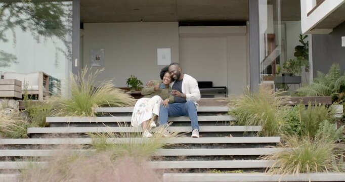 African American couple sitting on front steps, woman showing smartphone, man holding cup, laughing