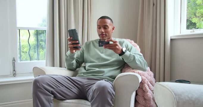 African American man in mint top sitting by window checking phone comparing card verifying order
