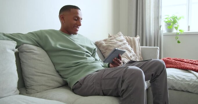 African American man leaning on sofa at home holding tablet and comparing small card with device