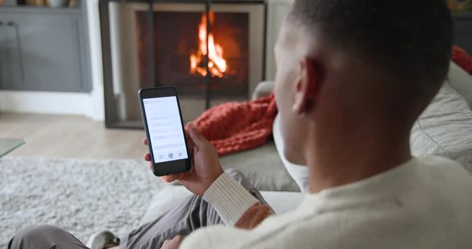 African American man sitting on right sofa with phone, reading, glancing at fireplace, copy space