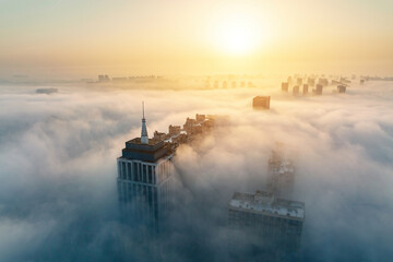 Fototapeta premium Tall urban buildings emerge from dense morning fog under the glow of a rising sun, creating a misty cityscape.