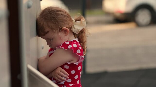 A sad little girl in a red dress with polka dots is standing on the street, hugging her shoulders, expressing loneliness, shyness, and childlike emotions.