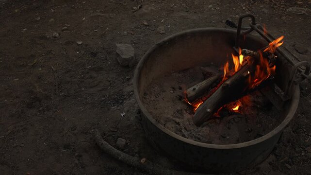 Burning logs create bright flames inside metal fire pit outdoors, glowing over ash and charred wood at a rustic campsite surrounded by bare ground