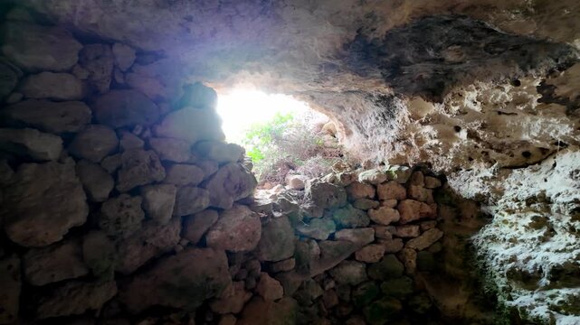 Light floods cave mouth, revealing ancient stone walls. Sunbeams create a halo effect through rocky archway. Texture of rough stones contrasts with natural cave ceiling. Scene suggests hidden history
