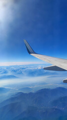 Fototapeta premium Airplane wing flying high above majestic snow-capped mountain ranges and layer of mist under a clear blue sky. View looking out of a passenger plane window during a sunny flight