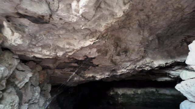 Dark cave interior with rough stone walls and ceiling. Glowing green algae cover water surface below. Light source casts eerie shadows across jagged rock formations