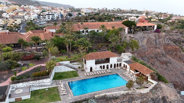Aerial view of a resort pool area with sun loungers, palm trees, and traditional architecture with terracotta roofs, situated on a cliffside in La Gomera, Canary Islands, Spain.