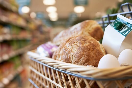 Close-up of a wicker shopping basket filled with fresh bread, milk, and eggs inside a grocery store aisle.