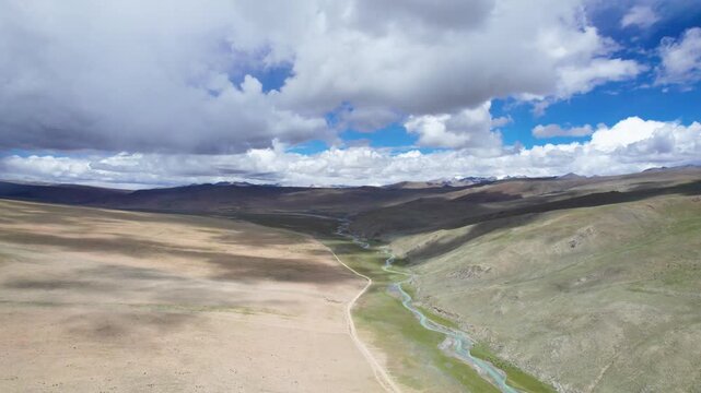 Drone view landscape of zigzag stream and off road path on Tibet plateau with shadows of clouds moving on desert land, blue sky white clouds and mountains,  4k real time footage b roll travel concept.