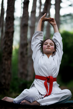 Young girl in a karate gi with a red belt meditating and stretching in a forest setting during martial arts practice