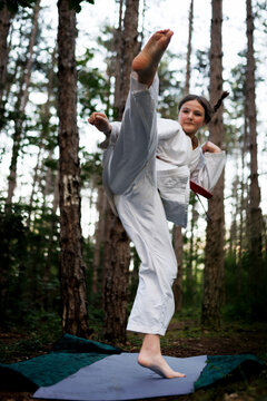 Young girl practicing karate high kick in forest clearing wearing a white gi and red belt showing strength and focus outdoors