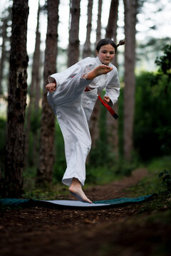 Young girl practicing a high karate kick outdoors in a pine forest wearing a white gi and red belt during training