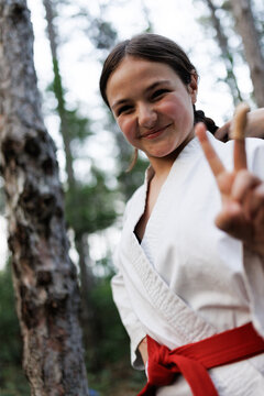 Smiling girl in a white karate gi with a red belt flashes a peace sign outdoors in a forest during martial arts training and practice