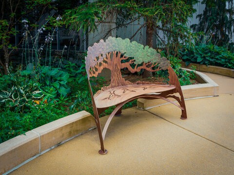 Unique metal tree design bench sculpture positioned outdoors in a scenic green garden plaza with stone paving