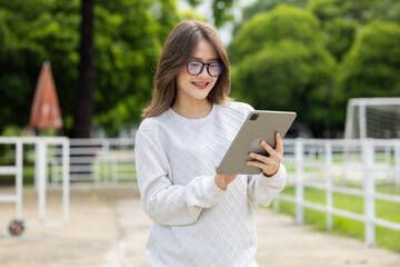Young woman tablet outdoor park eyeglasses sweater smile digital technology leisure standing in green park while using tablet with relaxed expression near white fence and tree background © 220 Selfmade studio
