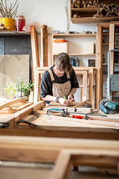 Concentrated female carpenter working in wood shop on antique wooden window frame restoration, craftswoman holding part of frame and marking wood with special pencil tool, preparing patch replacement.