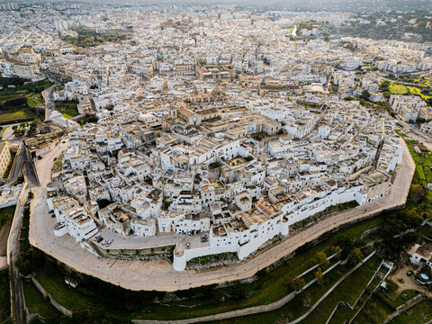 Aerial view of the historic white-washed buildings and narrow streets of the famous White City, surrounded by ancient defensive walls and lush countryside in Ostuni, Apulia, Italy.